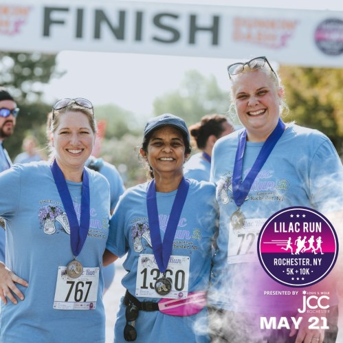 Three women in blue shirts smile at the camera, celebrating their finish at the Lilac Run in Rochester, NY.