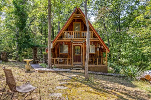 A charming A-frame cabin nestled among trees, featuring a wooden exterior and a small pathway leading to the entrance.