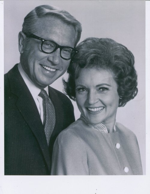 A smiling couple poses closely together, both dressed in vintage attire, with the man wearing glasses and the woman in a stylish coat.