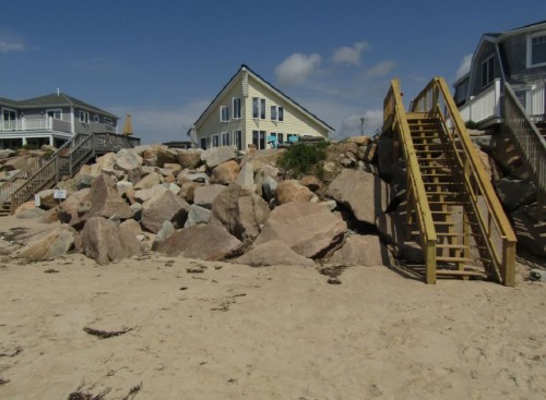 A sandy beach with large rocks and a wooden staircase leading to a house in the background under a blue sky.