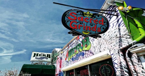 Colorful exterior of Sacred Grinds café with a vibrant mural and signage against a blue sky.
