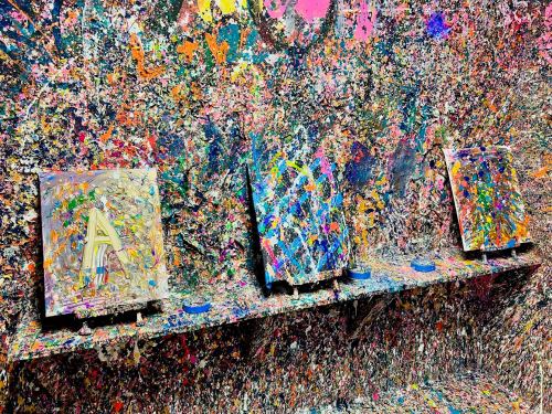 Three colorful canvases on a splattered wall, showcasing vibrant paint drips and abstract designs.