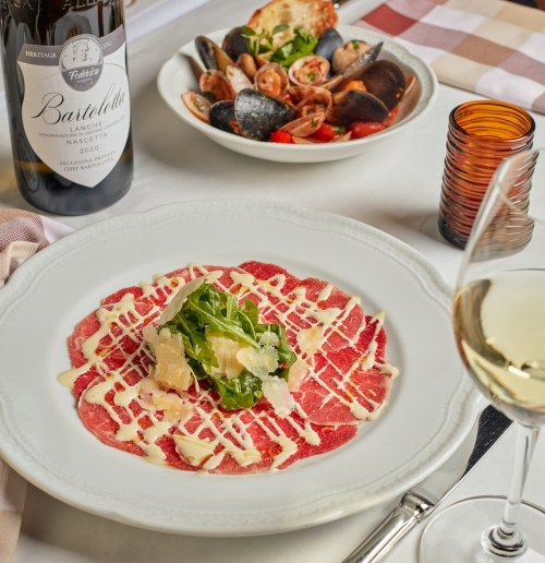 A plate of beef carpaccio drizzled with sauce, garnished with arugula, alongside a seafood dish and a bottle of wine.