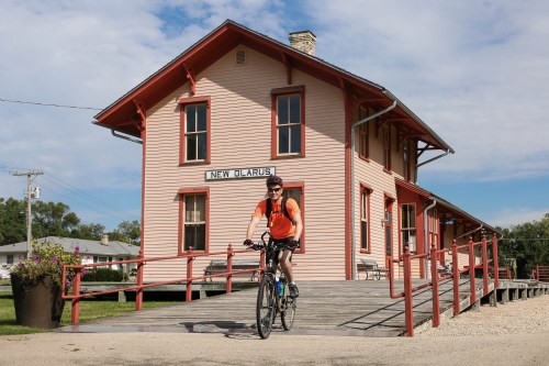 A cyclist rides past a historic pink train station labeled "New Glarus" under a blue sky with scattered clouds.