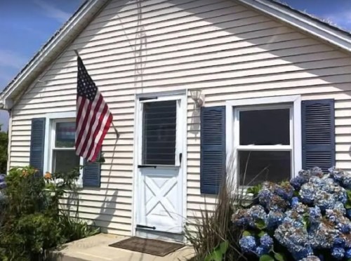 A charming house with a front door, blue shutters, an American flag, and blooming hydrangeas in the garden.