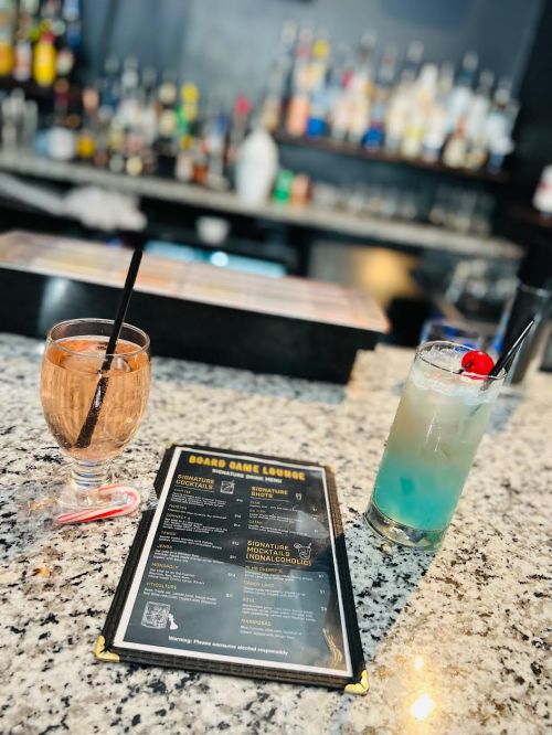 Two colorful cocktails sit on a granite bar top next to a menu for a board game lounge. Bottles are visible in the background.