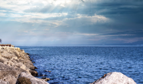 A serene view of a rocky shoreline meeting a calm blue sea under a cloudy sky.