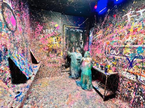 Two people in protective gear stand in a colorful, paint-splattered room with vibrant walls and scattered paint supplies.