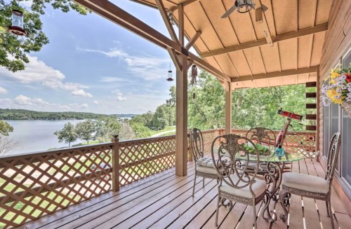 A spacious porch with a dining table overlooking a serene lake and lush greenery under a clear blue sky.