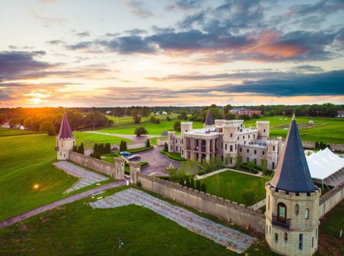 Aerial view of a castle with turrets, surrounded by green fields and a sunset in the background.