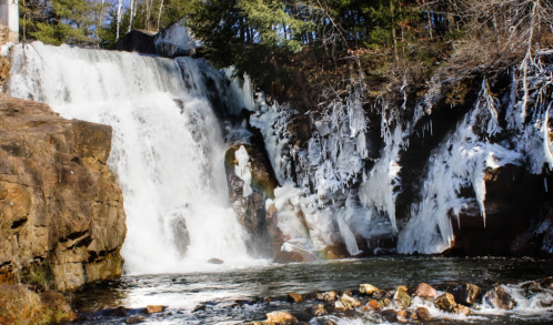 A frozen waterfall cascading over rocky cliffs, surrounded by trees and ice formations.