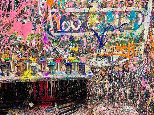 A vibrant wall splattered with colorful paint, with paint cans lined up on a table in front.