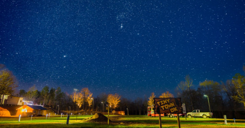 A starry night sky over a small town, with trees and buildings illuminated by soft lights. A sign reads "Tiny Town OK."
