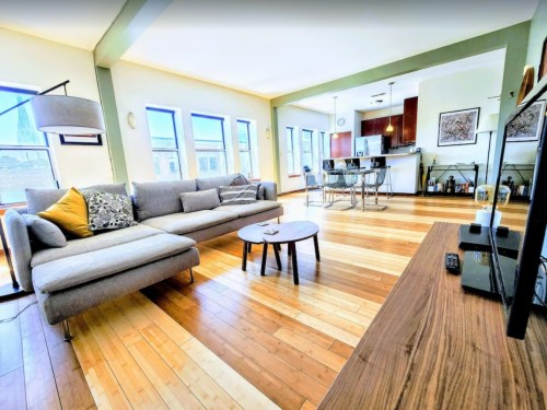 Bright living room with a gray sofa, wooden floor, and a view of a kitchen in the background. Cozy and modern decor.