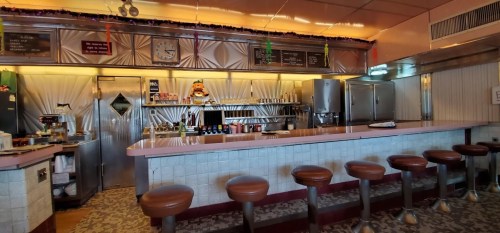 A retro diner interior featuring a long pink counter, bar stools, and vintage decor with festive decorations.
