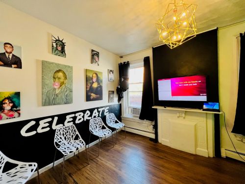 A cozy room with art on the walls, a TV, and white chairs, featuring the word "CELEBRATE" in bold on the wall.