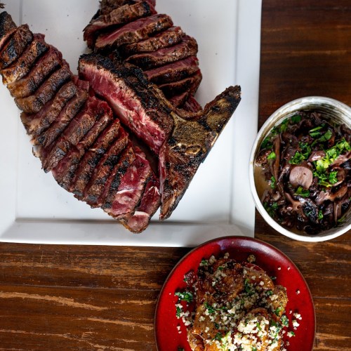 A platter of sliced grilled steak, accompanied by a bowl of beans and a small plate of seasoned grains.
