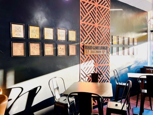 A cozy board game lounge with framed artwork on the wall and empty tables and chairs bathed in warm sunlight.