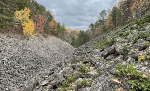 A rocky landscape with autumn foliage, featuring a steep, sloped valley under a cloudy sky.