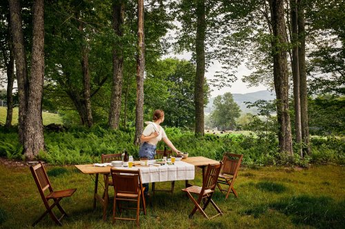 A woman sets a rustic table in a forest clearing, surrounded by trees and a scenic view of hills in the background.