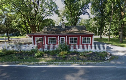 A charming red cottage with white trim and a picket fence, surrounded by greenery and trees along a quiet road.