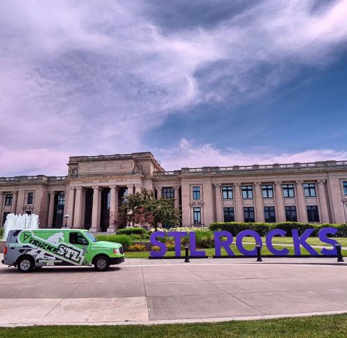 A green van parked near a large "STL ROCKS" sign in front of a historic building under a blue sky.