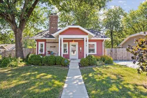 A charming red house with a front porch, surrounded by greenery and a well-maintained lawn on a sunny day.