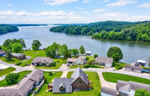 Aerial view of a serene lakeside community with houses, green trees, and calm water under a blue sky.