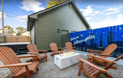 A cozy outdoor seating area with wooden chairs around a fire pit, set against a colorful "Welcome to Indianapolis" mural.