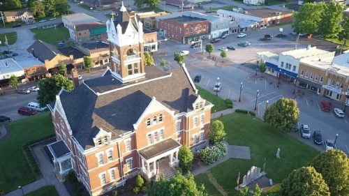 Aerial view of a historic brick courthouse surrounded by greenery and a small town with shops and streets in the background.