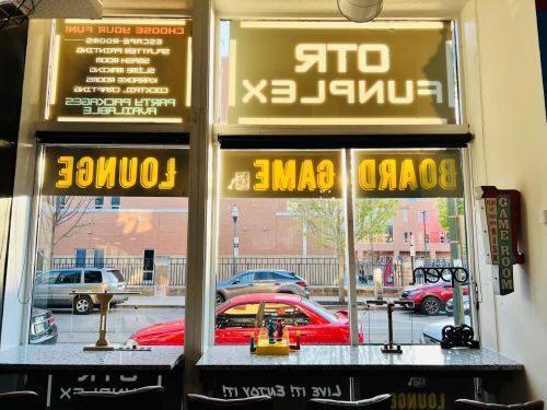 View from inside a game lounge with large windows displaying signs for board games and a red car outside.