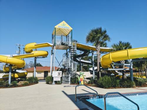 A bright yellow water slide structure with multiple slides, surrounded by palm trees and a clear blue sky.