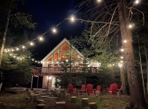 A cozy cabin at night, illuminated by string lights, surrounded by trees and featuring a fire pit and red chairs.