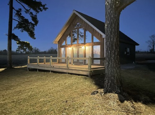 A cozy cabin with large windows and a deck, illuminated at night, surrounded by trees and open fields.