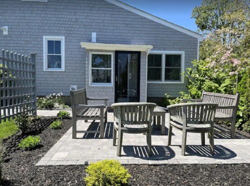 A cozy outdoor seating area with wooden chairs and a table, surrounded by greenery and a light-colored house.