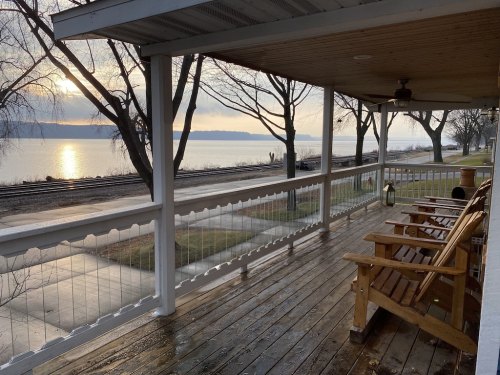A peaceful porch view overlooking a lake at sunset, with wooden chairs and trees lining the shore.
