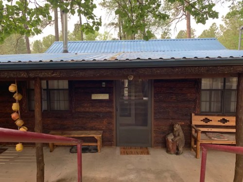 A rustic log cabin with a blue metal roof, featuring a front porch and wooden benches, surrounded by trees.