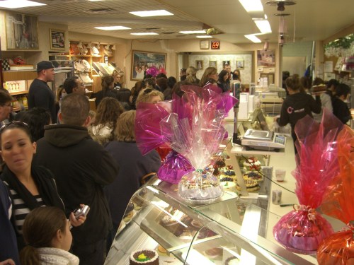 A busy bakery with a long line of customers, colorful gift wraps on display, and a variety of pastries in the foreground.