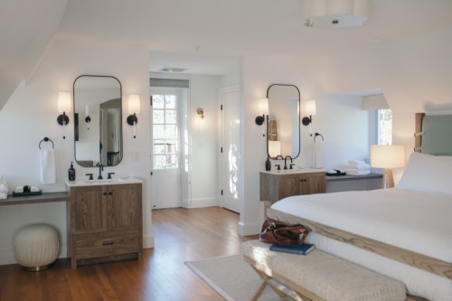 Bright, modern bedroom with a large bed, wooden furniture, and two mirrors, leading to a light-filled bathroom area.