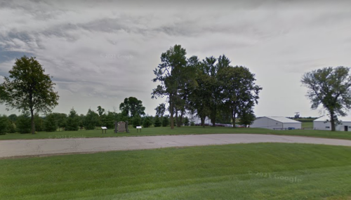 A grassy area with trees and a gravel road, featuring a stone monument and a building in the background.