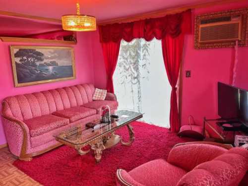 A vibrant pink living room featuring a plush sofa, glass coffee table, and red curtains, with a painting on the wall.