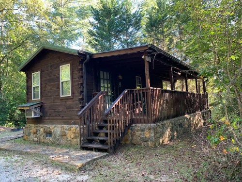 A rustic wooden cabin with a stone foundation, surrounded by trees and featuring a front porch and steps.
