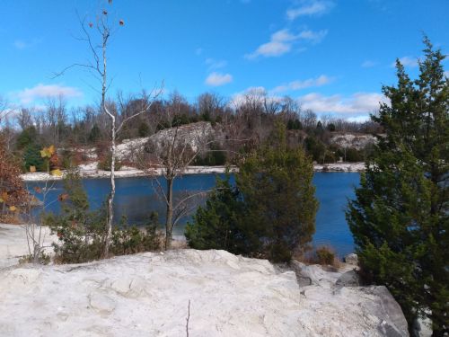 A serene lakeside scene with rocky shores, trees, and a clear blue sky dotted with clouds.