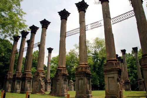 Ruins of tall, weathered columns stand in a green landscape under a cloudy sky, surrounded by trees.