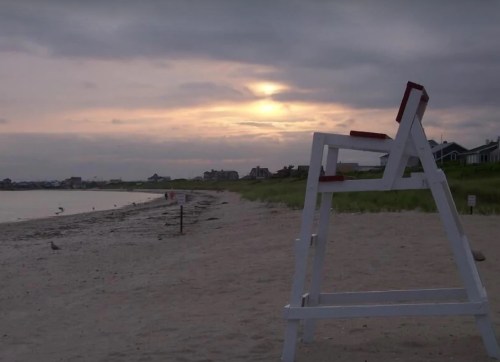 A lifeguard chair on a sandy beach at sunset, with a cloudy sky and distant houses along the shore.