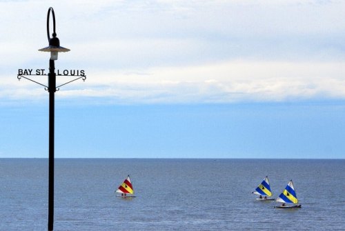 Sailboats with colorful sails on calm waters near a signpost for Bay St. Louis under a clear blue sky.