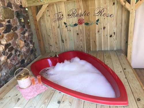 A heart-shaped red bathtub filled with bubbles, set in a wooden cabin with "The Rustic Rose" sign above.