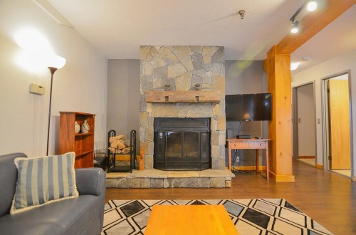 Cozy living room featuring a stone fireplace, wooden furniture, and a TV, with warm lighting and a patterned rug.