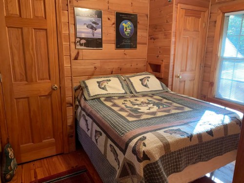 Cozy wooden bedroom with a double bed, quilted cover, and nature-themed posters on the walls. Sunlight streams in through a window.