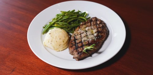 A plate featuring a grilled steak, mashed potatoes, and green beans, garnished with a sprig of rosemary.